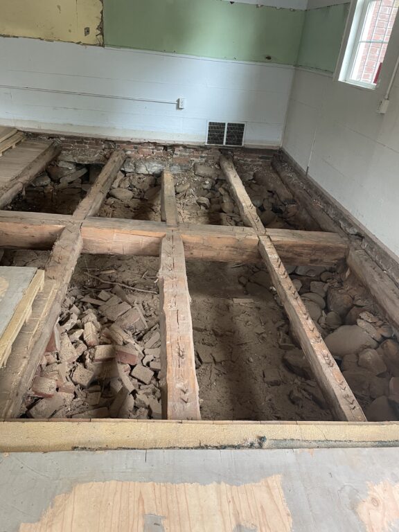 Interior view of a school room under renovation with exposed floor joists and debris, showing ongoing construction work.